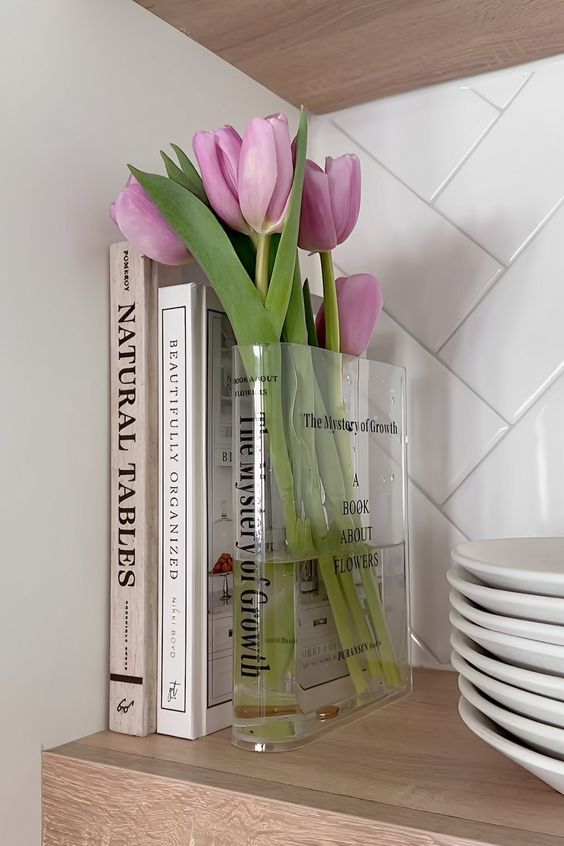 pink tulips are in a clear vase next to books on a wooden shelf