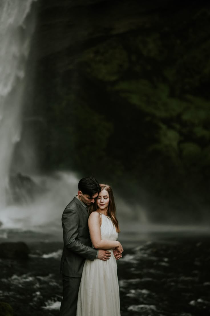 a bride and groom standing in front of a waterfall with their arms around each other