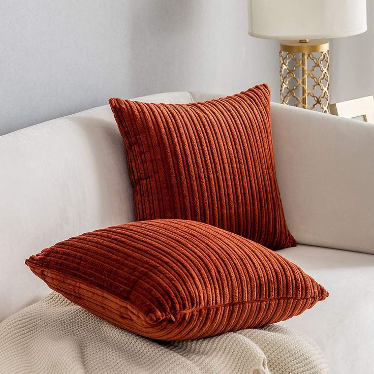 two orange pillows sitting on top of a white couch next to a lamp and window