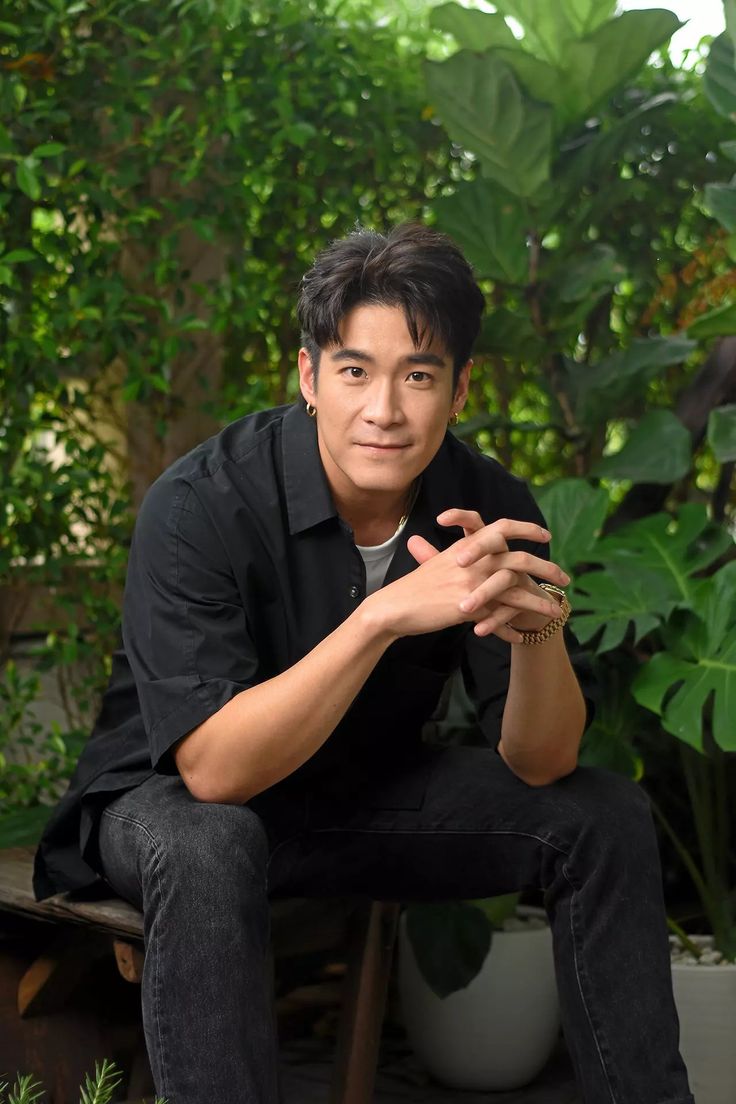 a man sitting on top of a wooden bench in front of some trees and plants