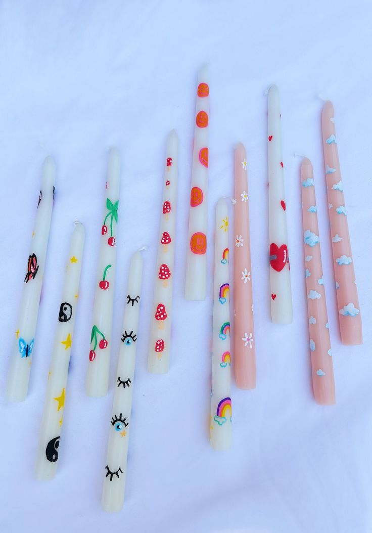 five different colored candles are lined up on a white tablecloth, with designs painted on them