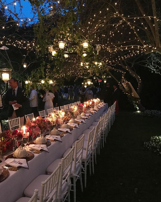 a long table is set up with white chairs and lights strung from the trees in the background