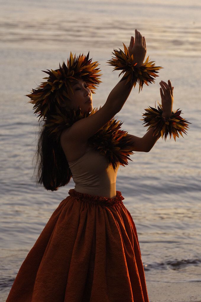 a woman in an orange skirt holding two large flowers up to her face while standing on the beach