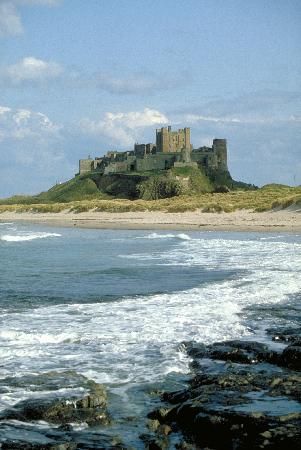 a castle sitting on top of a green hill next to the ocean with waves crashing in front of it