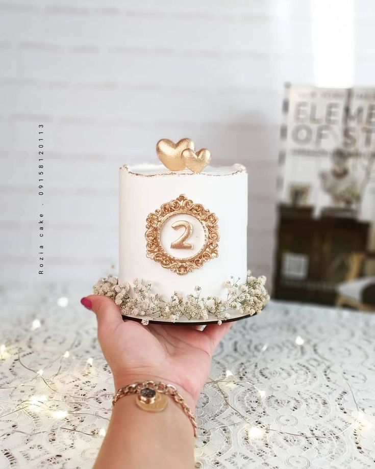 a hand holding a white cake with gold decorations on it and the number two on top