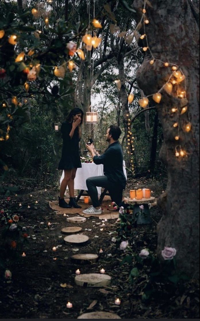 a man and woman sitting at a table in the middle of a forest surrounded by candles