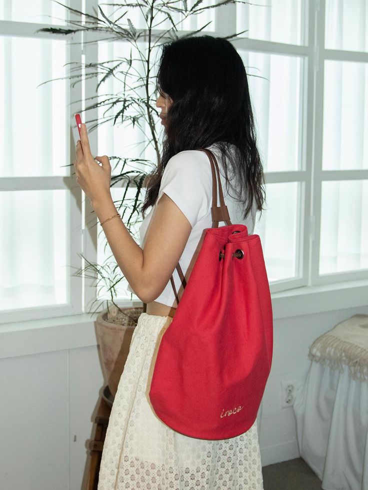 a woman standing in front of a window with a red bag on her shoulder and cell phone in her hand