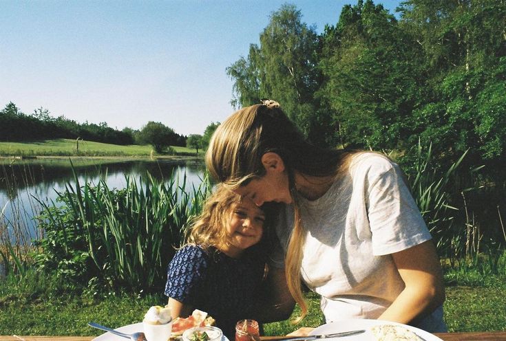 a woman and child sitting at a picnic table with food in front of the water