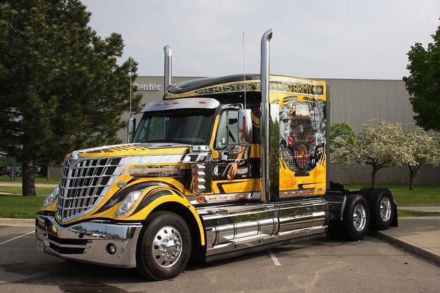 a large yellow semi truck parked in a parking lot next to trees and building behind it