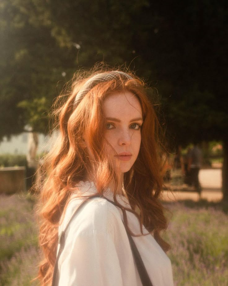 a woman with long red hair is standing in a field and looking at the camera