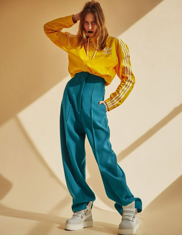 a woman in yellow shirt and teal pants posing for the camera with her hands on her head