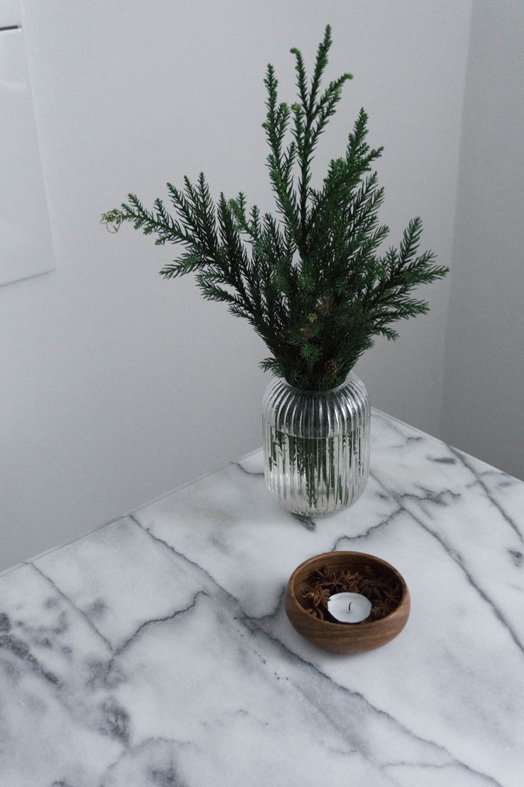 a white marble table with a glass vase filled with greenery and a wooden bowl