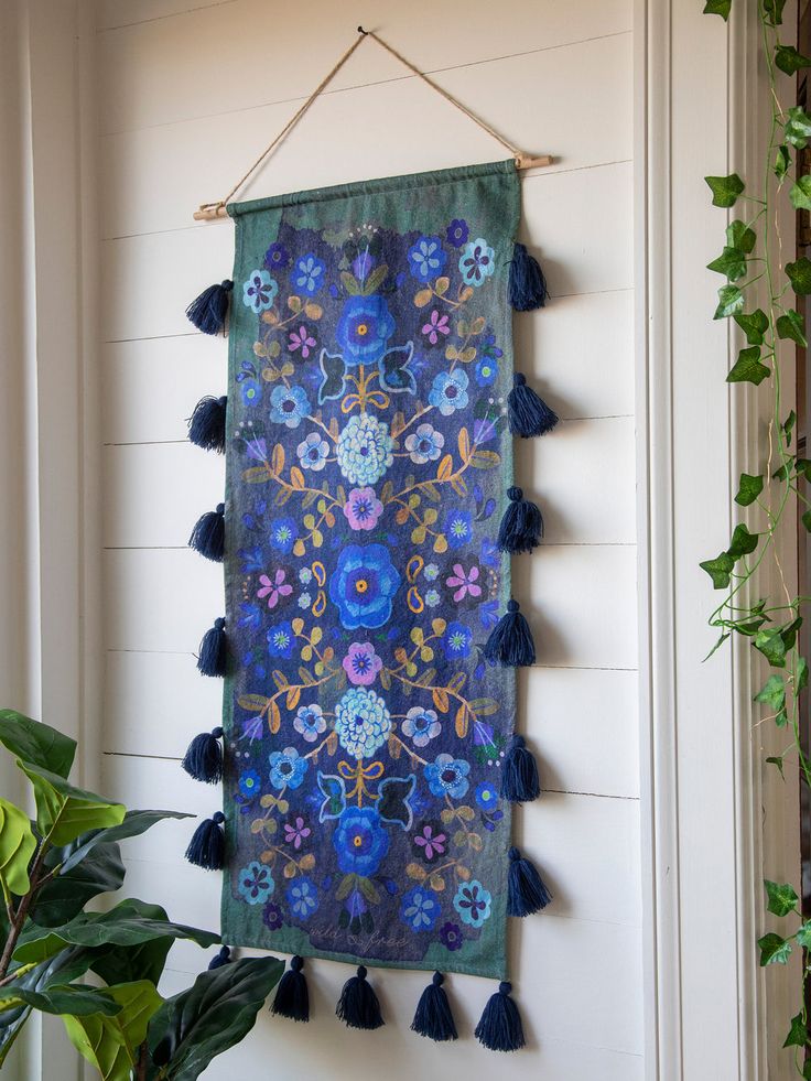 a blue wall hanging with tassels and flowers on the side of a house