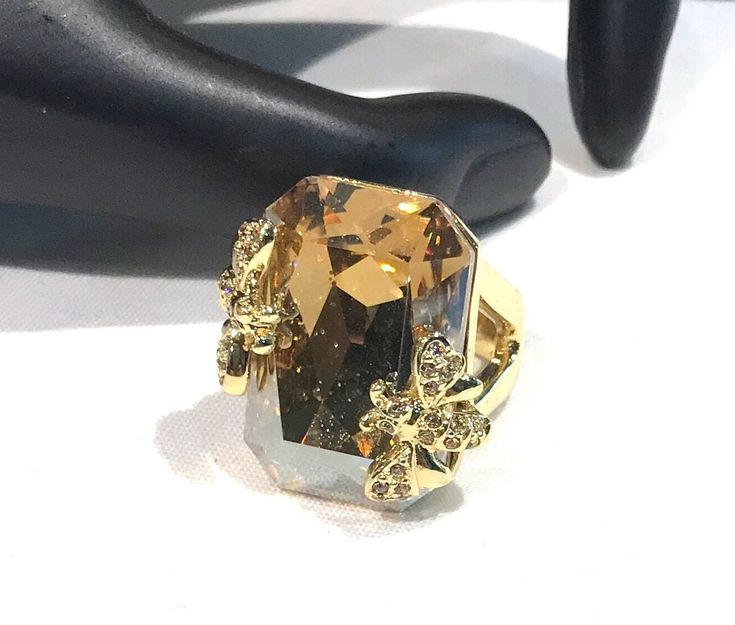 This gorgeous vintage  sparkly statement ring features a large rectangular shaped champagne coloured  faceted crystal with raised  pave  rhinestone flying insects on each side of the stone.  The sides of the ring has a triangle shape open work and  is a size 6.50 and the face 1" x .75" and is in very good/excellent condition with the champagne crystal excellent looking  very good/excellent and the gold tone great.  Unsigned and weighs 22 grams Shipping prices vary on the weight of item. I do my Glamorous Gold Crystal Ring For Party, Gold Crystal Ring With Bling For Party, Gold Bling Crystal Ring For Party, Gold Rectangular Crystal Ring, Elegant Gold Rectangular Crystal Ring, Gold Crystal Ring With Diamond Accents For Party, Gold Crystal Ring With Rhinestones For Party, Gold Crystal Ring With Rhinestones For Formal Occasions, Luxury Gold Crystal Party Ring