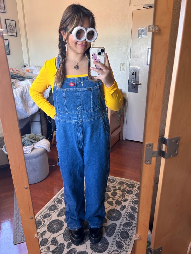 a woman wearing overalls taking a selfie with her cell phone in the mirror