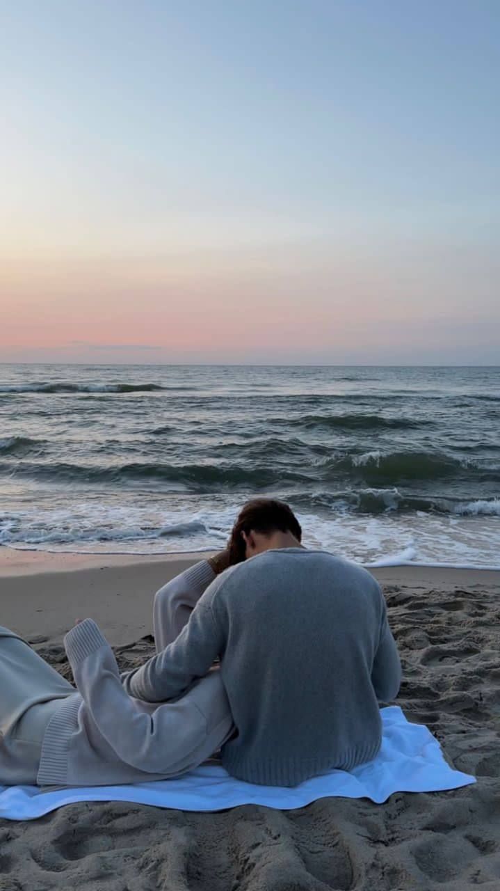 a woman sitting on top of a blanket on the beach next to the ocean at sunset