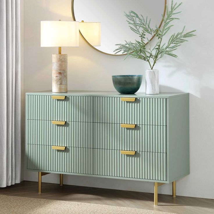 a green dresser with gold handles and drawers in front of a round mirror on the wall