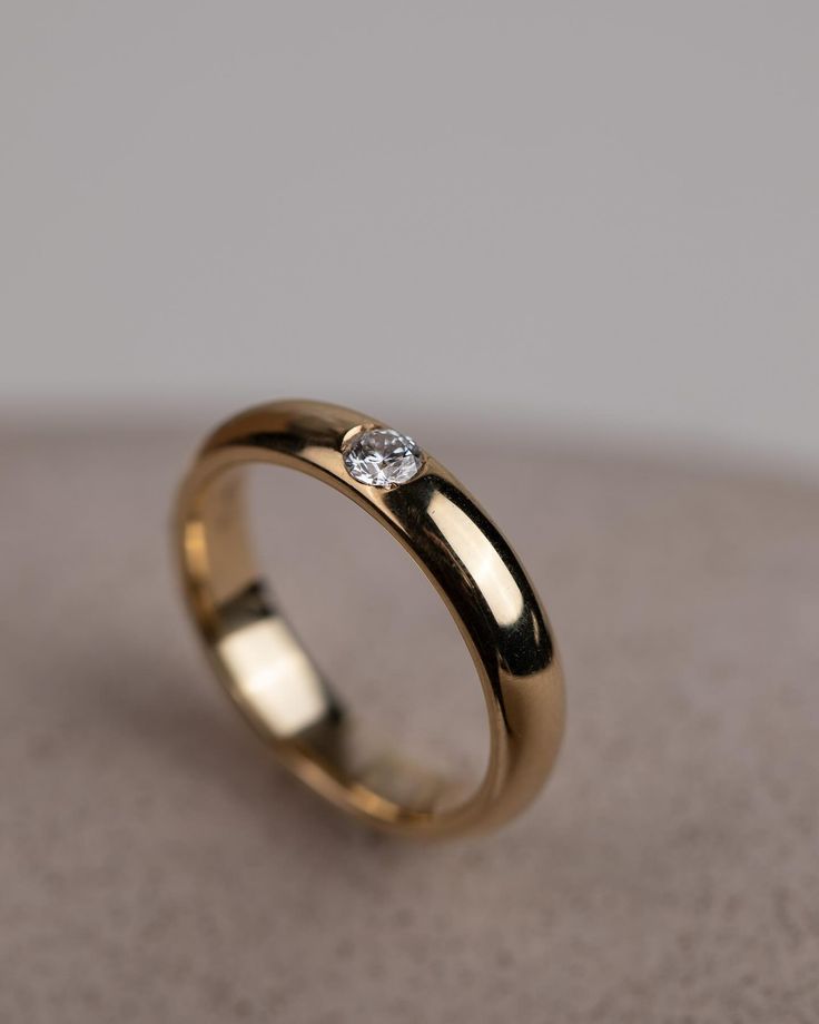 a gold wedding ring with a single diamond in the center on a beige surface,