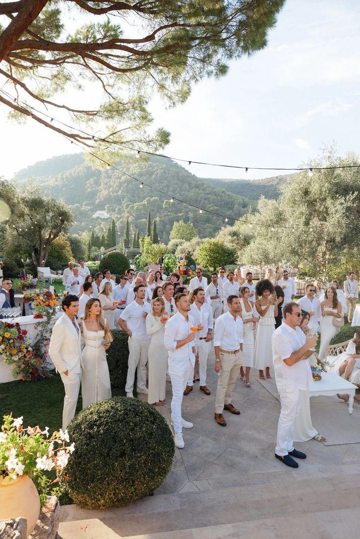 a group of people standing around each other in front of some trees and bushes on a sunny day