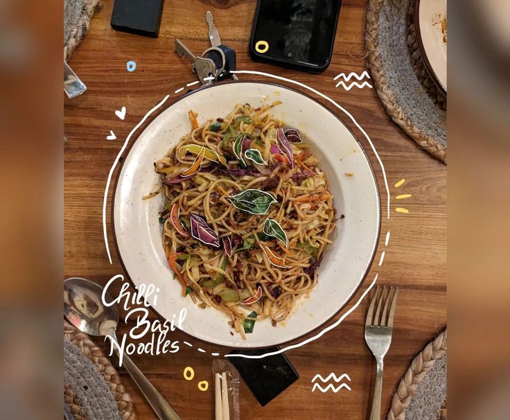 a white plate topped with pasta next to utensils and phone on top of a wooden table