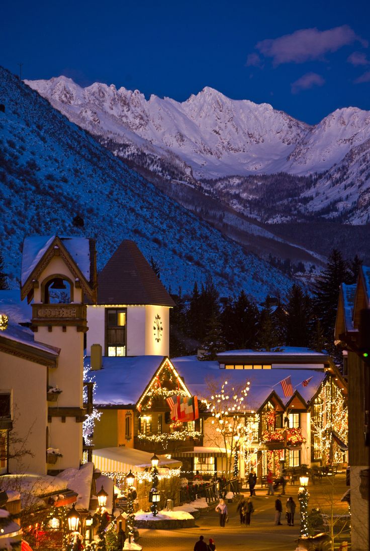 the town is lit up with christmas lights and snow covered mountains in the background at night