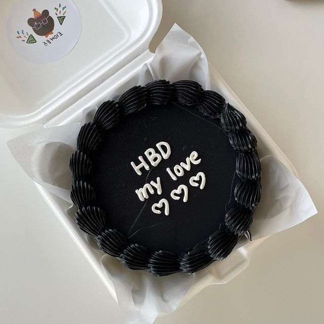 a black cake with writing on it sitting next to a plastic container filled with fruit