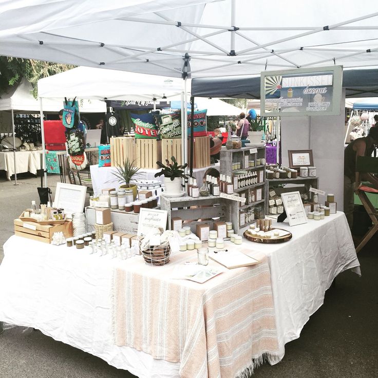 an outdoor market with many items on the table and people standing around it under a white tent