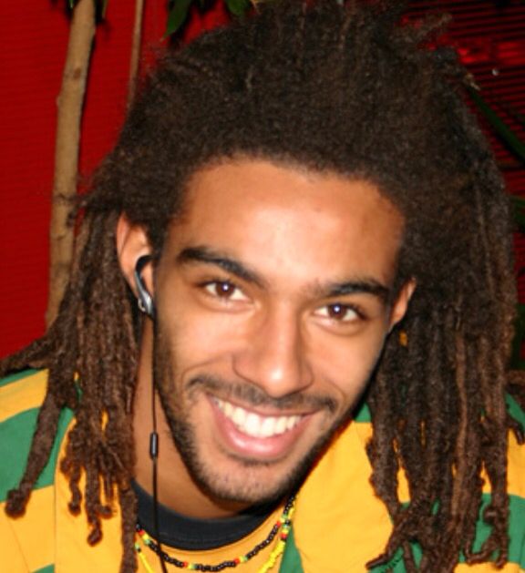 a man with dreadlocks smiles at the camera while wearing a green and yellow striped shirt