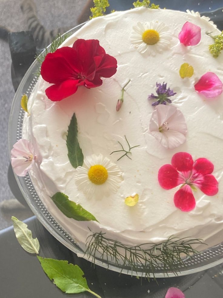 a white cake with flowers on it sitting on a glass table next to some leaves