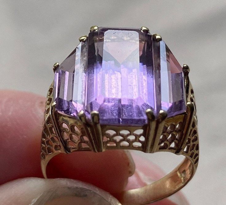 "14 karat yellow gold and amethyst ring.  The delicate filigree setting features three amethysts.  The center stone measures 9/16\" long and 5/8\" wide. It is an emerald cut amethyst flanked by 2 trapezoid cut amethysts.  The stones are of a light/medium violet color.  This is a statement ring in a simple symmetrical style.  Size is approximately 5." Art Deco Amethyst Ring In Yellow Gold, Yellow Gold Amethyst Ring In Art Deco Style, Art Deco Yellow Gold Amethyst Ring, 14k Gold Hallmarked Purple Amethyst Ring, Hallmarked 14k Gold Purple Amethyst Ring, Purple Amethyst Ring Stamped 14k, Purple Amethyst Ring With Intricate Design, Purple Hallmarked Art Deco Amethyst Ring, Art Deco Purple Amethyst Ring