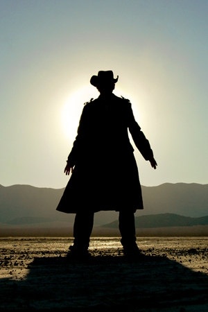 the silhouette of a man in a long coat and cowboy hat standing on a beach