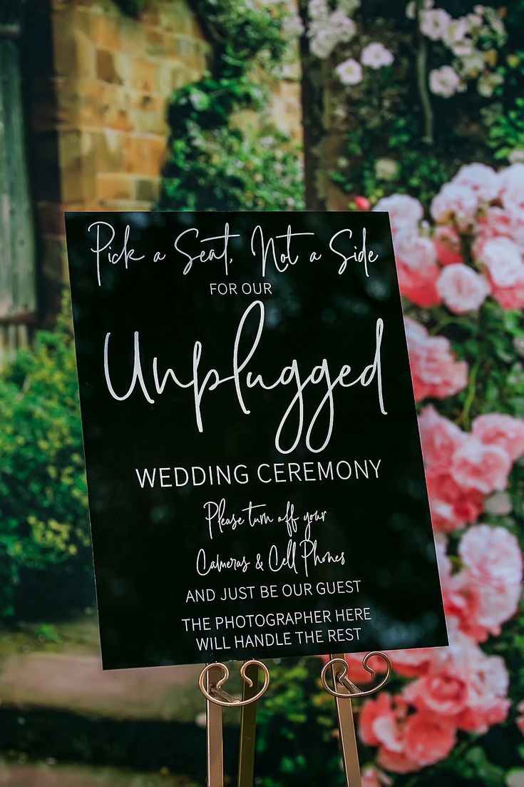 a sign that is standing up in front of a flower garden with pink flowers behind it