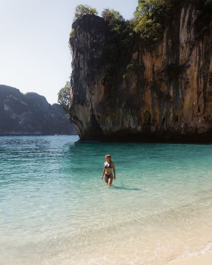 a woman is wading in the clear blue water near an island with large cliffs