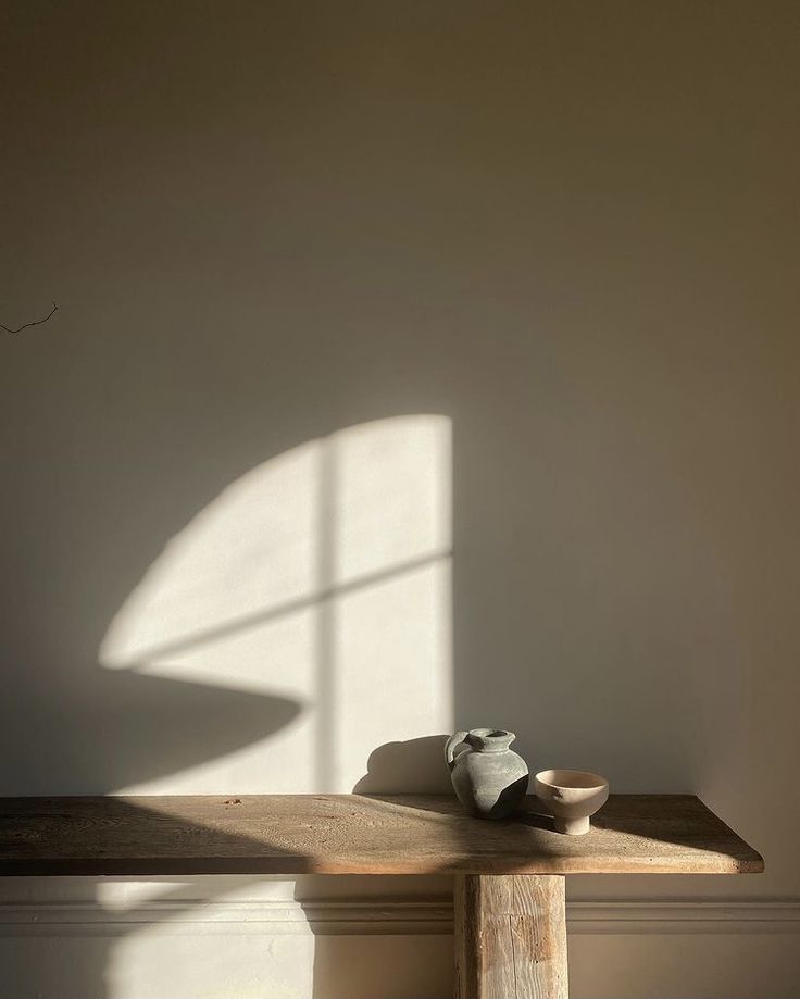 a wooden table with a vase and bowl on it next to a window in a white room