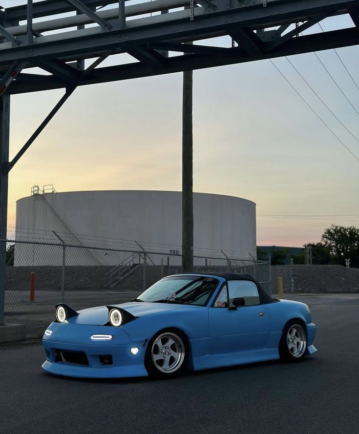 a blue sports car parked in front of a gas tank at sunset or dawn with its hood down