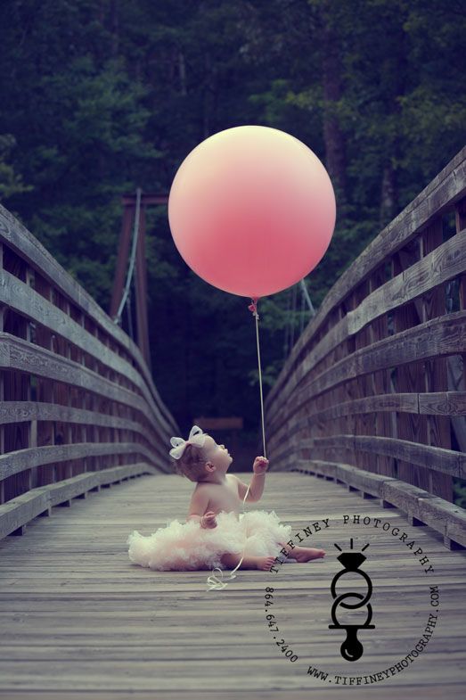 a teddy bear holding a pink balloon while sitting on top of a wooden bridge with trees in the background