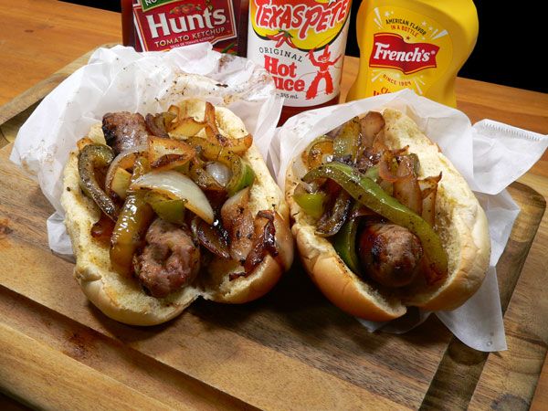 two hot dogs with onions, peppers and mustard on a cutting board next to condiments