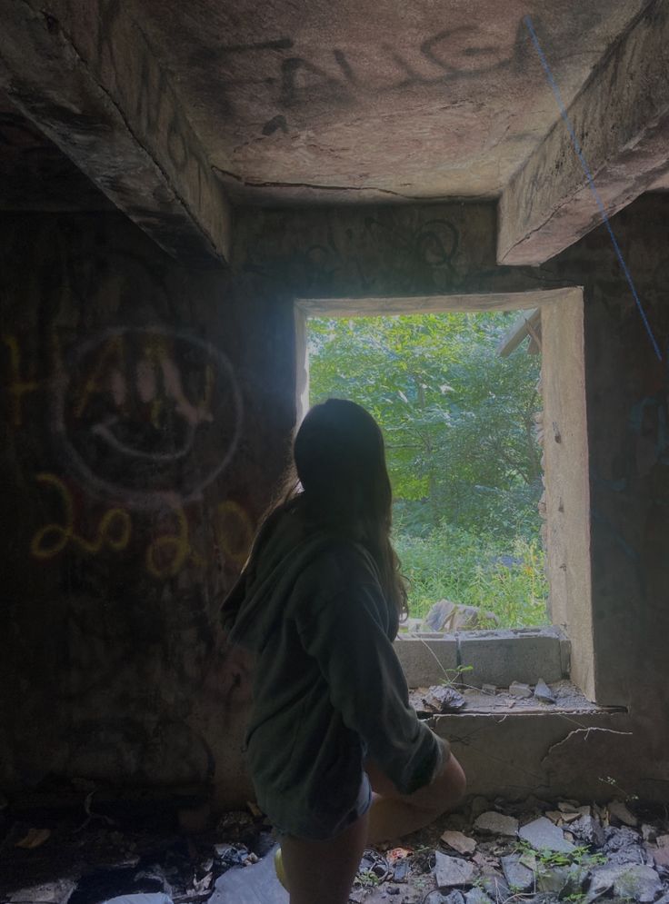 a person standing in an abandoned room with graffiti on the walls and windows, looking out