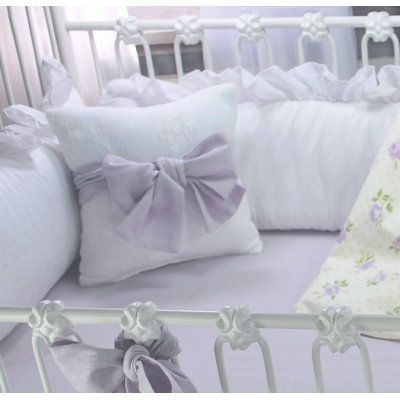 a baby crib with white sheets and purple bows on the bedding, next to pillows