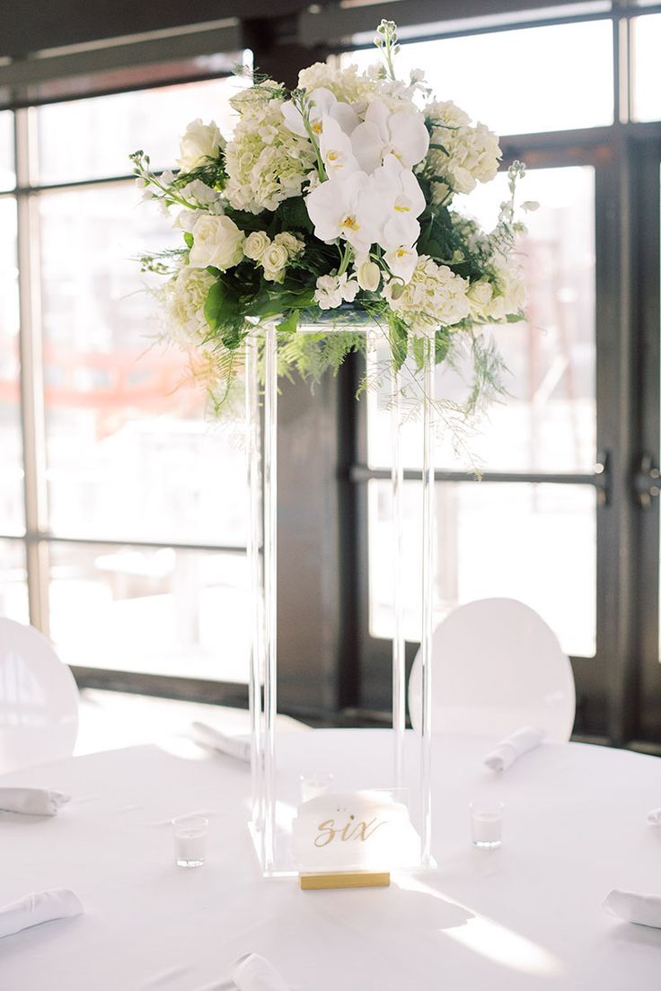 a tall vase with white flowers and greenery sits on top of a round table