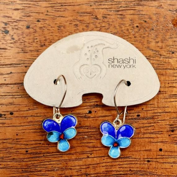 This graceful, Art Nouveau inspired design shows a pansy flower in bloom. Handcrafted of 24k gold over sterling silver. The enameled earrings are offered in hinged french backs for pierced ears. Size: 1 1/4 inches.The Shashi earrings offered here are vintage stock from the 1970's and 1980's. They were designed by the Shashi Singapuri, crafted by hand, and were part of a line featured on the cover of vogue (quite a handful of times) and numerous other fashion magazines. Gold Enamel Earrings With Flower Charm, Gold Earrings With Flower Charm In Enamel, Gold Earrings With Flower Charm And Enamel, Blue Enamel Flower Earrings As Gift, Enamel Flower Charm Earrings, Enamel Flower Earrings With Flower Charm, Blue Flower Hoop Earrings Gift, Blue Flower Hoop Earrings As Gift, Blue Flower-shaped Hoop Earrings