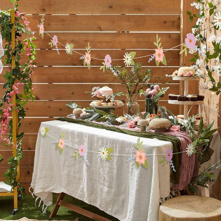 a table with flowers and cakes on it in front of a wooden fence, surrounded by greenery