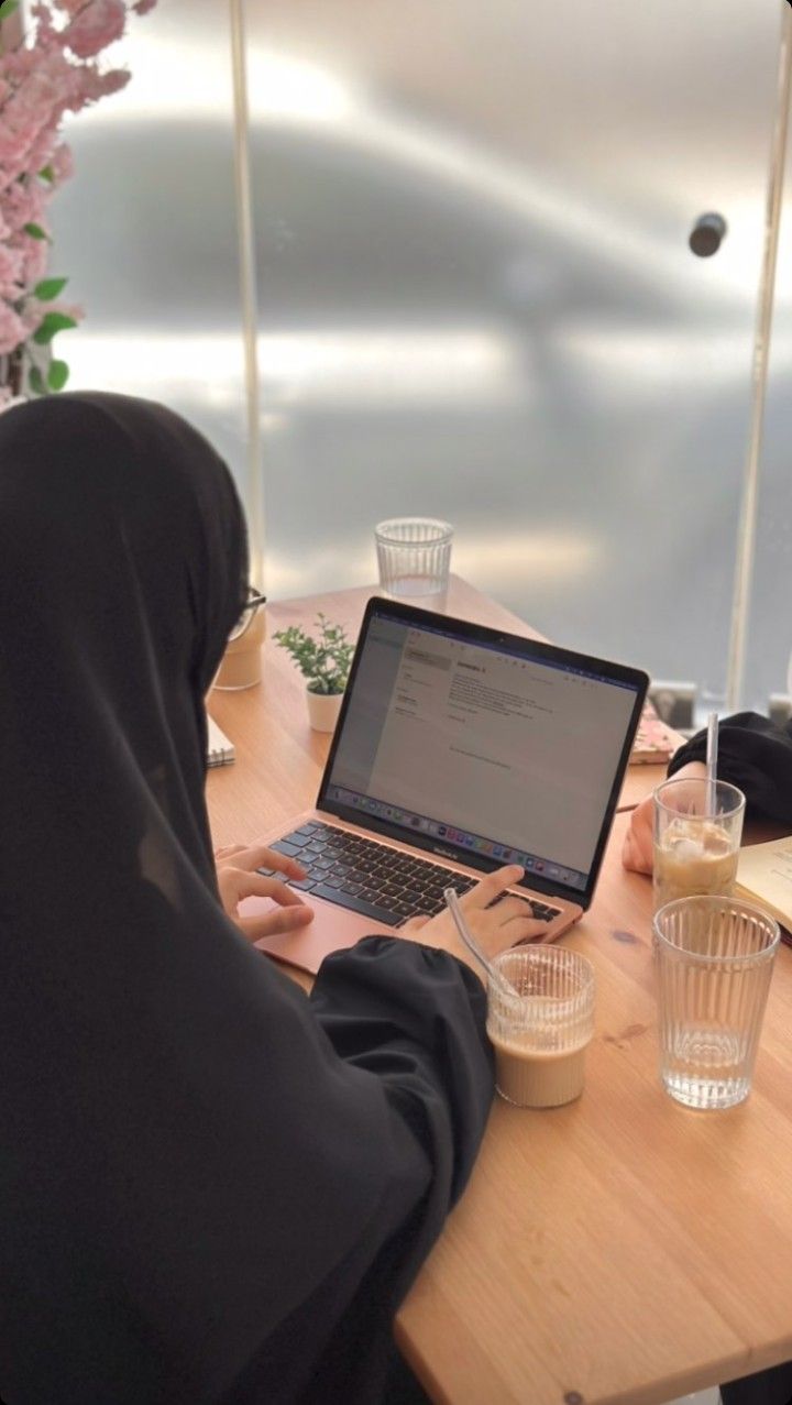 a woman sitting at a table with a laptop computer in front of her, wearing a hijab