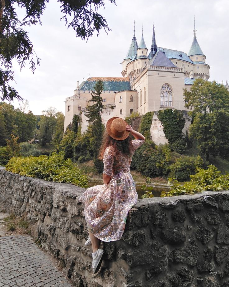 a woman in a dress and hat sitting on a stone wall looking at a castle