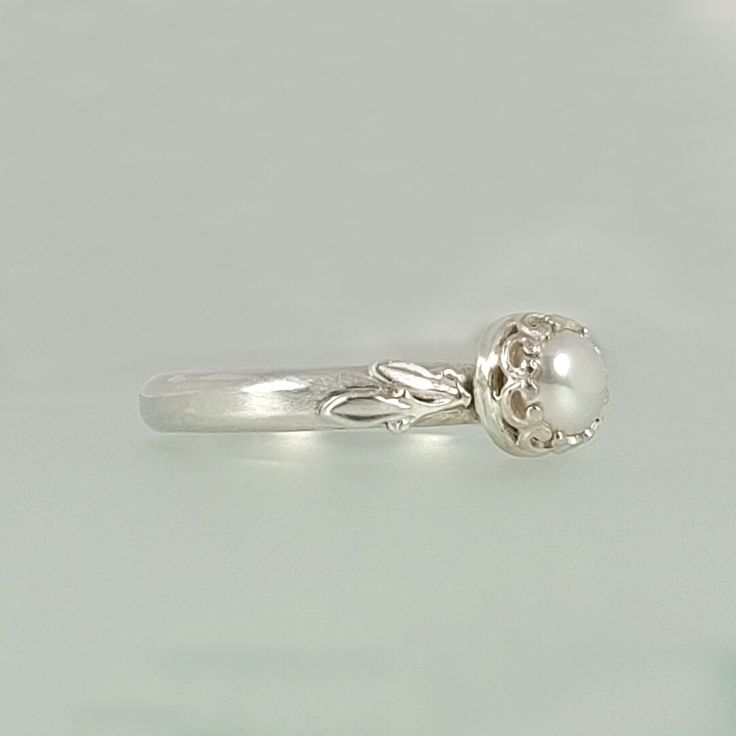 a silver ring with a pearl on the top and two leaves on the bottom, sitting on a white surface