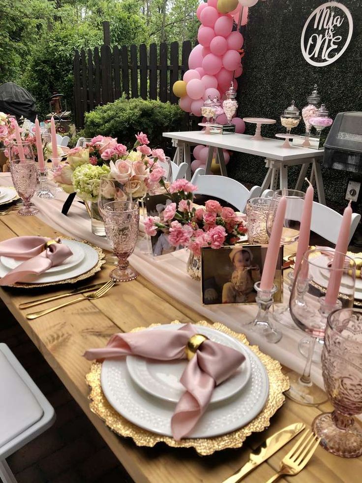 a table set with pink and white plates, silverware, napkins and balloons in the background