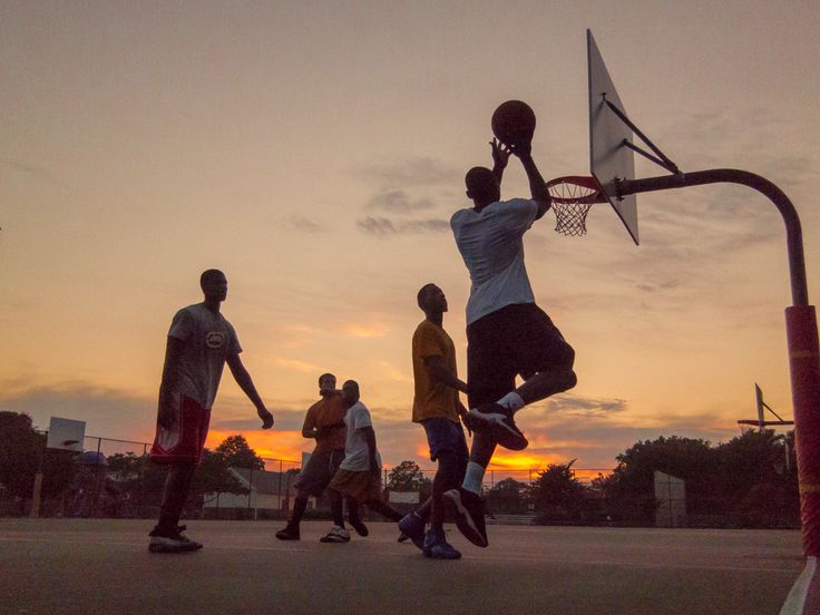 a group of young men playing basketball against each other on a court at sunset or dawn