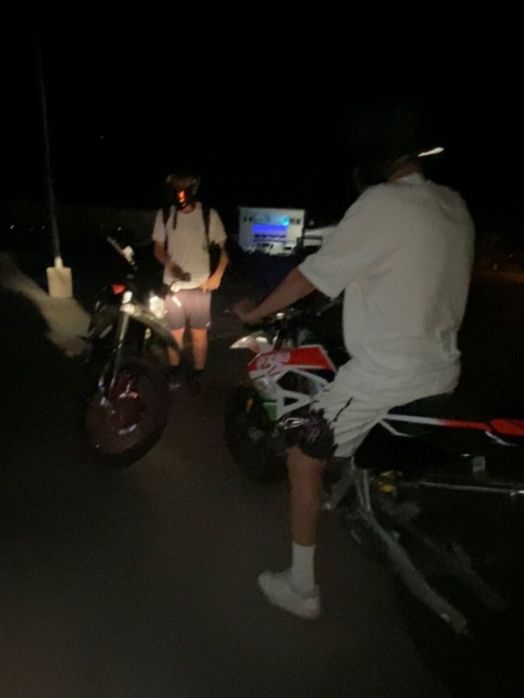 two people on motorbikes in the dark with one person standing next to them