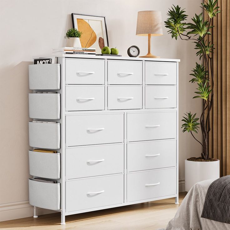 a white dresser with many drawers in a room next to a lamp and potted plant