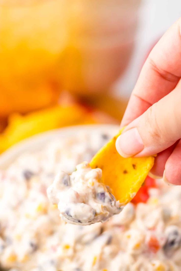 a hand holding a tortilla chip over a bowl of dip with cheese and black olives